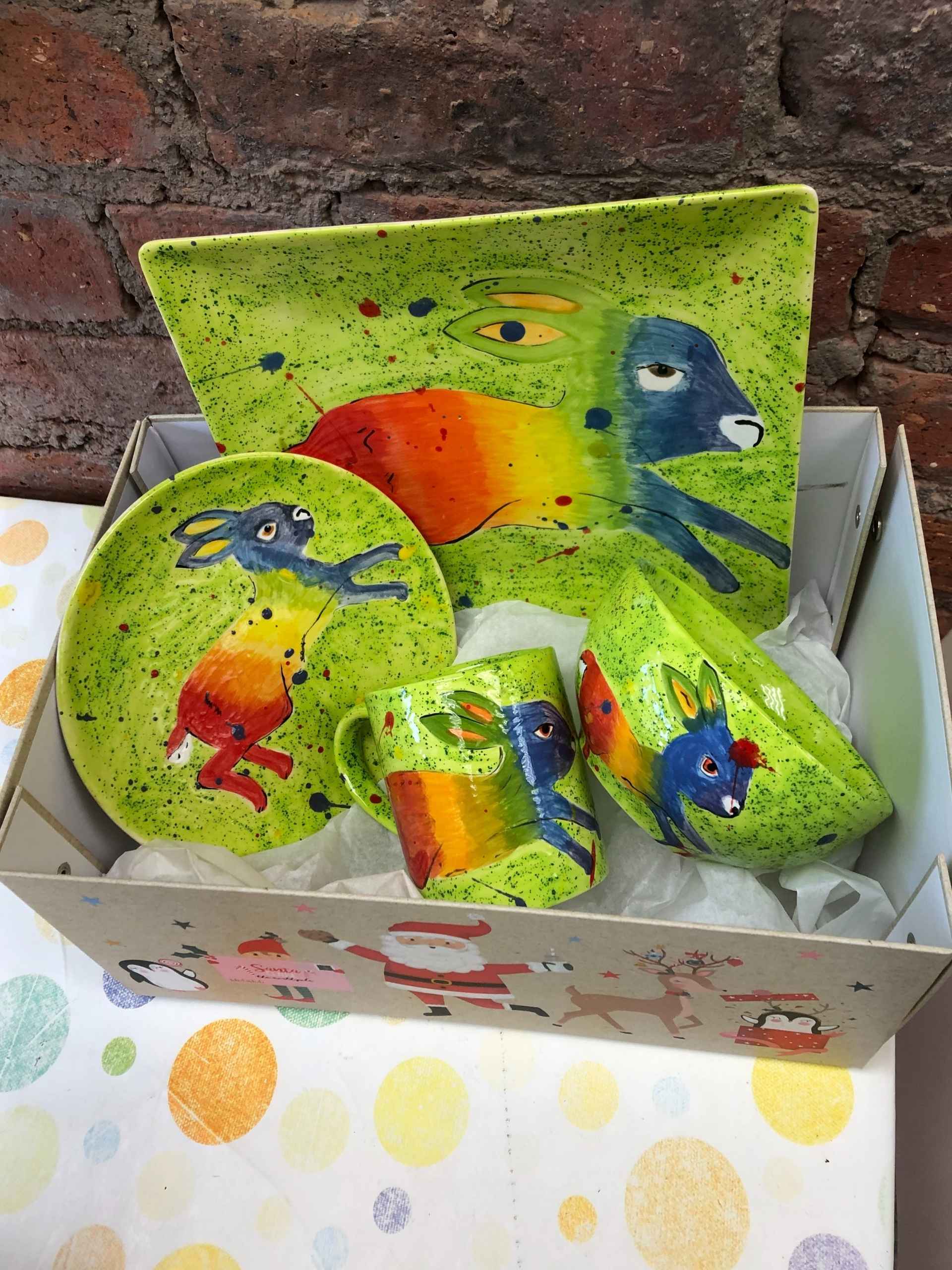 A box in a pottery studio containing two colourful plates, a bowl, and a cup, all featuring a rabbit design on a green background.