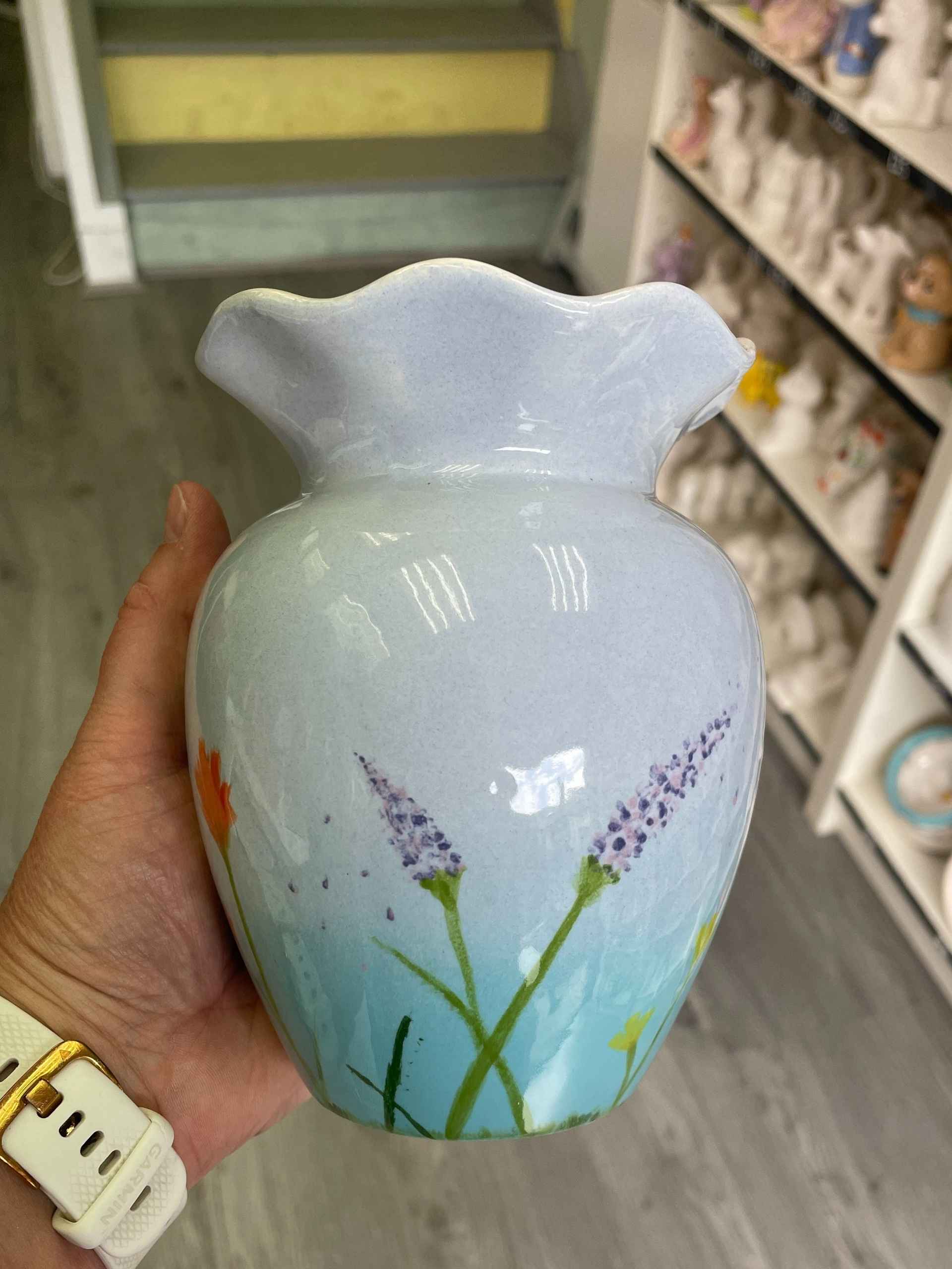 A hand gently holds a decorative vase adorned with vibrant flowers, showcasing a blend of colours and textures. Pottery studio In Hillsborough